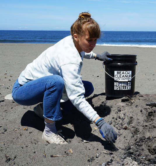 A woman picks up trash on a beach, while holding a bucket